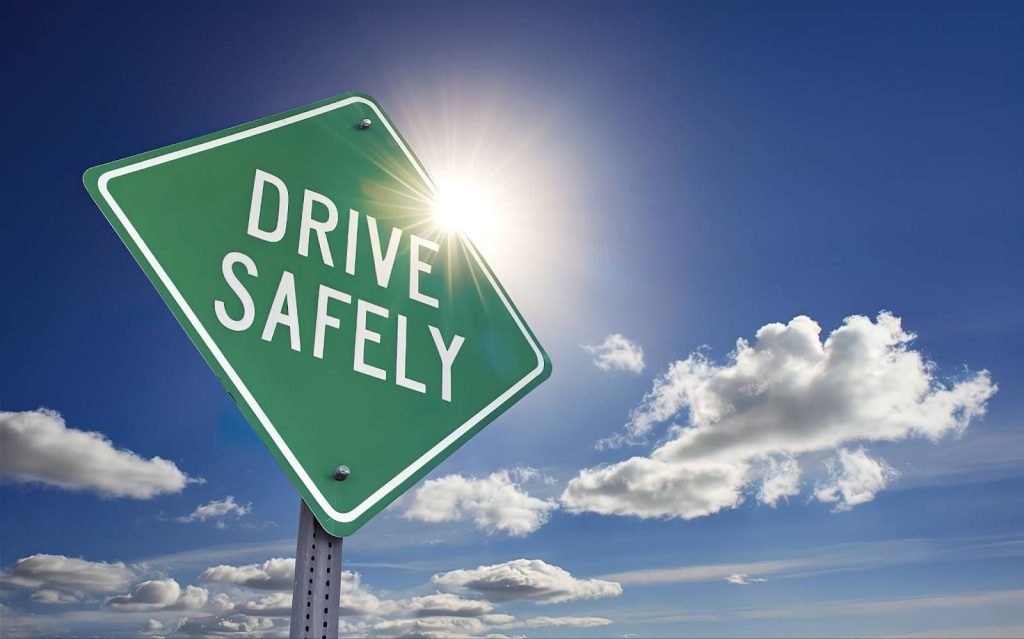 A green street sign that says “Drive Safely” standing against a blue sky.