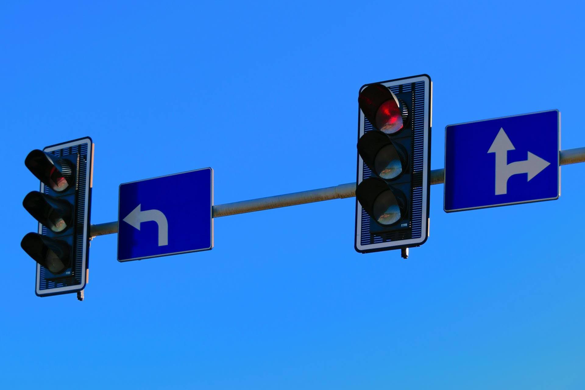 Traffic lights hanging over an intersection against a clear blue sky, with one signal showing red. Blue road signs display directional arrows