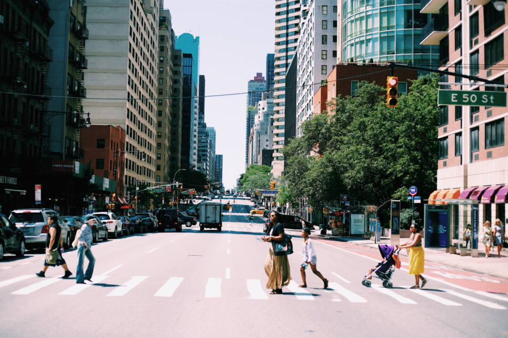 Pedestrians walk along a crosswalk on a street lined with tall buildings and cars. 