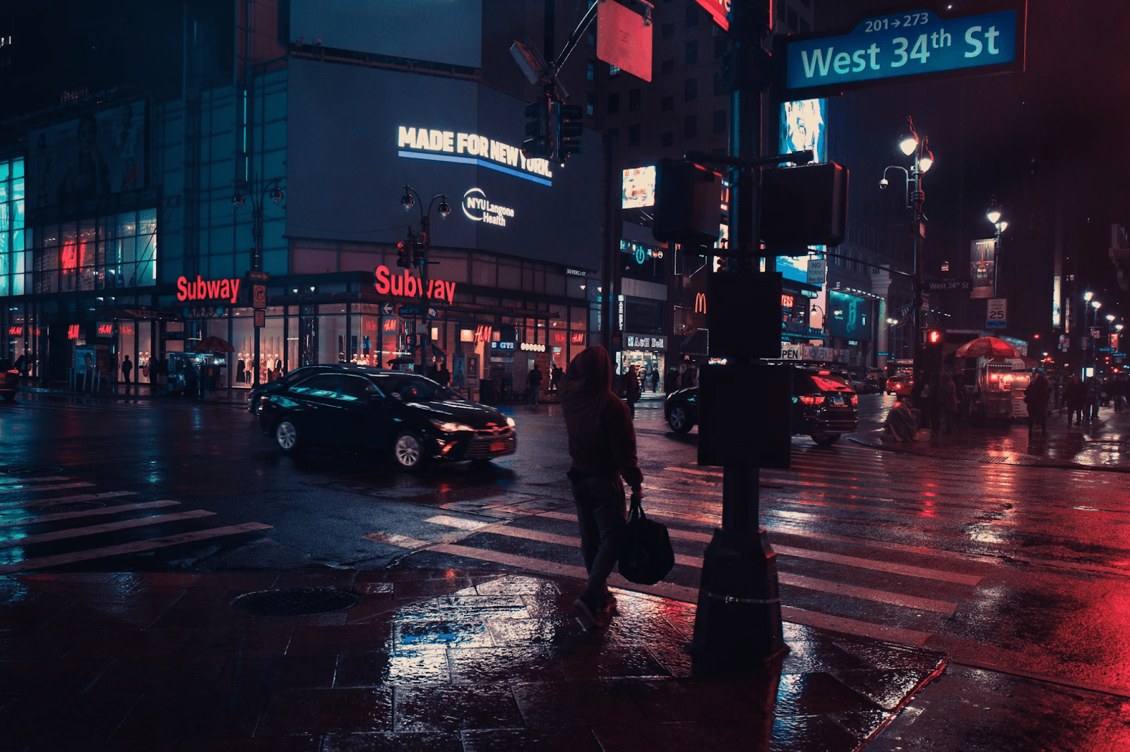 A wet intersection at night near an urban center with pedestrians and cars crossing through.